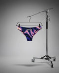 Purple and pink patterned thong on a hanger attached to a camera stand against a gray background