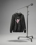 Black hoodie with pink graphic on a hanger next to a studio light stand against a gray background